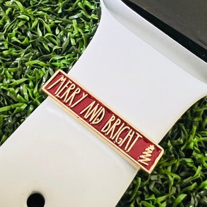 May include: A gold-toned watch band charm with the words "MERRY AND BRIGHT" in white lettering on a red background. A small Christmas tree design is on the right side. The charm is attached to a white watch band.