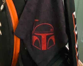 May include: A black golf towel with a red Boba Fett helmet graphic. The towel is hanging on a black golf bag.