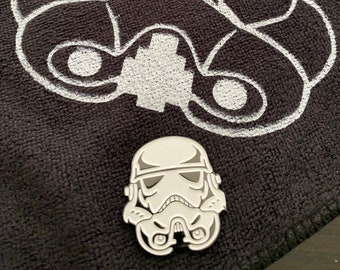 May include: A white enamel pin of a Star Wars Stormtrooper helmet. The pin is on a black fabric background with a larger white outline of a Stormtrooper helmet.