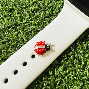 May include: A white smartwatch band with a red and black ladybug charm. The ladybug has silver accents and is attached to the band. The band is on a green surface.