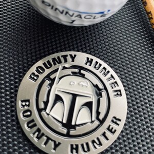 Puede incluir: Un marcador de bola de golf plateado con una imagen en blanco y negro del casco de Boba Fett. El marcador es circular y tiene las palabras "BOUNTY HUNTER" escritas alrededor del borde.