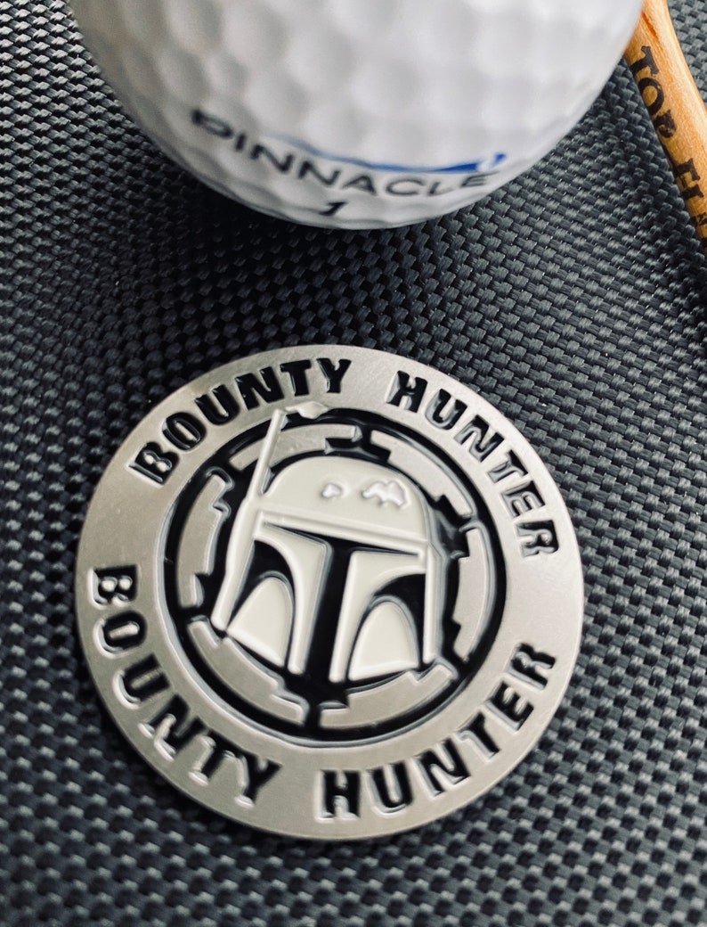 Puede incluir: Un marcador de bola de golf plateado con una imagen en blanco y negro del casco de Boba Fett. El marcador es circular y tiene las palabras "BOUNTY HUNTER" escritas alrededor del borde.