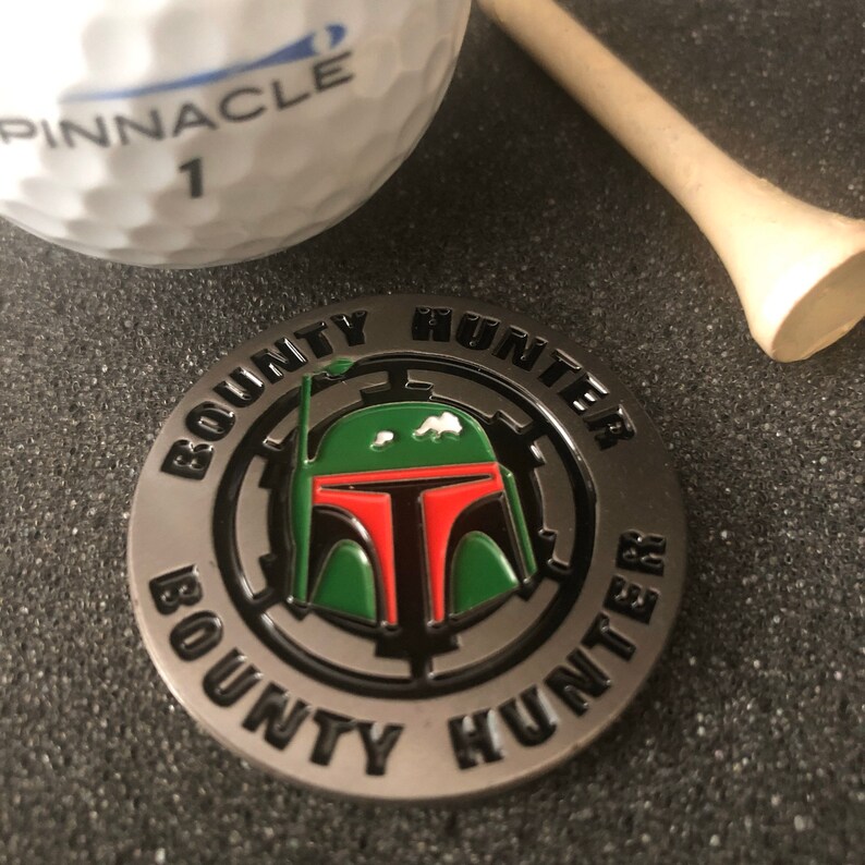 Puede incluir: Un marcador de bola de golf plateado con un dise&ntilde;o circular en blanco y negro. El centro del dise&ntilde;o presenta un casco Boba Fett verde y rojo con una visera negra. El texto "BOUNTY HUNTER" est&aacute; escrito alrededor del exterior del c&iacute;rculo.