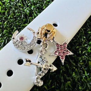 May include: A white watch band with a silver charm featuring a crescent moon, a star, and an arrow and bow. The charms are decorated with pink and clear rhinestones.