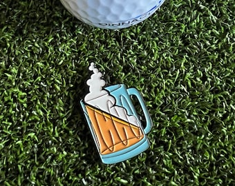 May include: A blue and yellow enamel pin of a beer mug with a foamy head. The pin is on a green artificial grass surface.