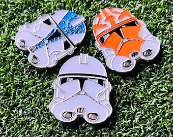 May include: Three Star Wars clone trooper helmet pins. The helmets are white with black details and have a silver finish. One helmet has a blue and white paint splatter design. Another helmet is orange with black details.