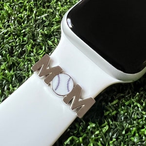 May include: A silver-toned watch band charm spelling "MOM" with a baseball design. The charm is attached to a white watch band. The background is a field of green artificial turf. The watch face is black.