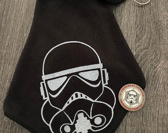 May include: Black golf towel with a silver Stormtrooper helmet graphic. Includes a silver metal clip and a round Imperial Forces coin. The towel is folded and has a loop for hanging.