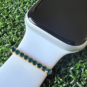 May include: A white Apple Watch band with a gold and teal blue crystal embellishment.
