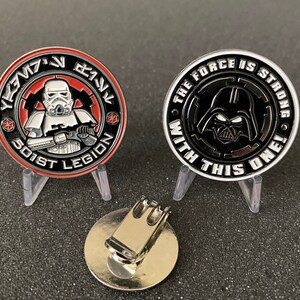 May include: Two collectible coins featuring Star Wars characters. One coin has a Stormtrooper and the text "501st Legion". The other coin has Darth Vader and the text "The Force is Strong With This One!". A silver-colored magnetic hat clip is also present.