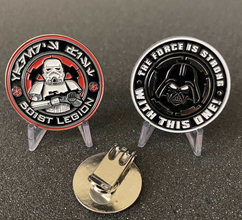May include: Two collectible coins featuring Star Wars characters. One coin has a Stormtrooper and the text "501st Legion". The other coin has Darth Vader and the text "The Force is Strong With This One!". A silver-colored magnetic hat clip is also present.