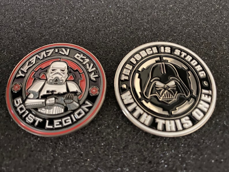 May include: Two collectible coins featuring Star Wars characters. One coin depicts a Stormtrooper with "501st Legion" text. The other coin shows Darth Vader with the text "The Force is Strong With This One!"