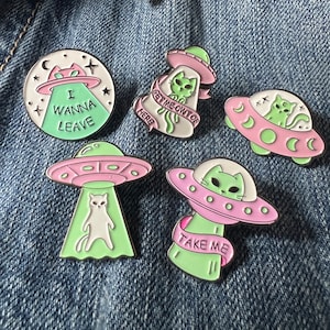 May include: Five enamel pins featuring green cats in pink and green spaceships. The pins say "I wanna leave", "Get me out of here", "Take me", and have moon and star designs.
