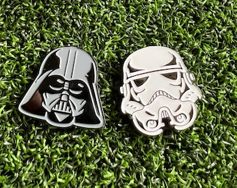 May include: Two enamel pins featuring Star Wars characters. One pin depicts Darth Vader's helmet in black and gray, and the other shows a Stormtrooper helmet in white and black. The pins are set against a green, textured background.