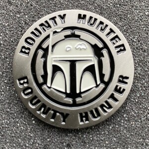 Puede incluir: Una moneda de metal de color plateado con una imagen en blanco y negro de un casco de cazarrecompensas de Star Wars. La moneda tiene las palabras "BOUNTY HUNTER" escritas alrededor del borde.