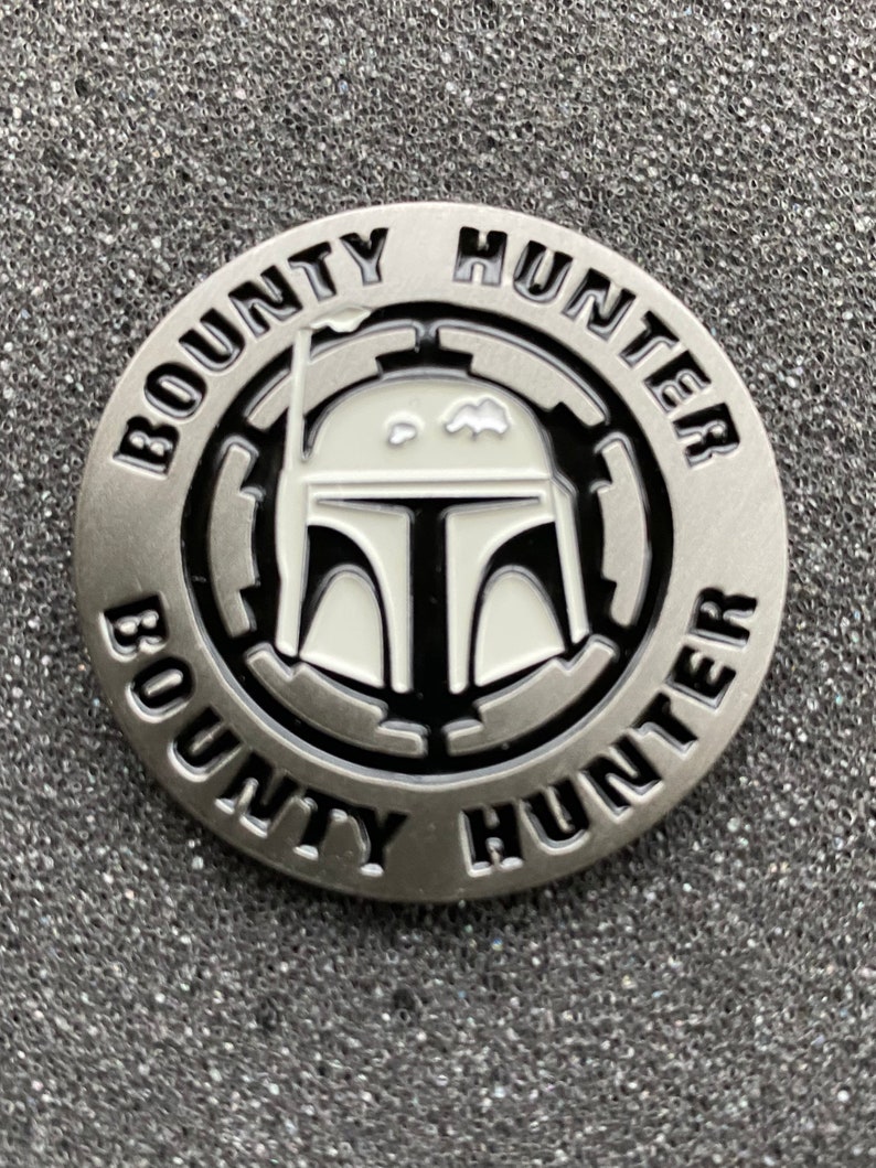 Puede incluir: Una moneda de metal de color plateado con una imagen en blanco y negro de un casco de cazarrecompensas de Star Wars. La moneda tiene las palabras "BOUNTY HUNTER" escritas alrededor del borde.