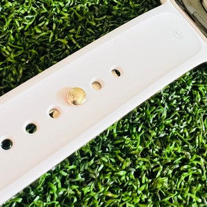 K&ouml;nnte beinhalten: Wei&szlig;es Apple Watch Armband mit einer goldenen Schlie&szlig;e und f&uuml;nf L&ouml;chern zum Einstellen der Gr&ouml;&szlig;e. Das Armband liegt auf einer gr&uuml;nen k&uuml;nstlichen Grasfl&auml;che.