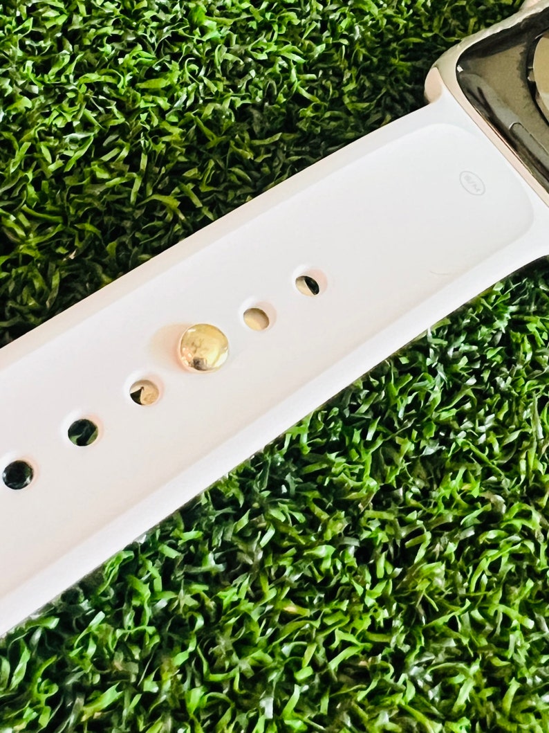K&ouml;nnte beinhalten: Wei&szlig;es Apple Watch Armband mit einer goldenen Schlie&szlig;e und f&uuml;nf L&ouml;chern zum Einstellen der Gr&ouml;&szlig;e. Das Armband liegt auf einer gr&uuml;nen k&uuml;nstlichen Grasfl&auml;che.