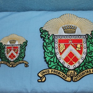 May include: Two embroidered patches featuring a crest with a red and white shield, a crown, and a sunburst. The larger patch has the text "Unus Probitas Omega Delta Sigma Integritas Ductus" on a gold scroll.