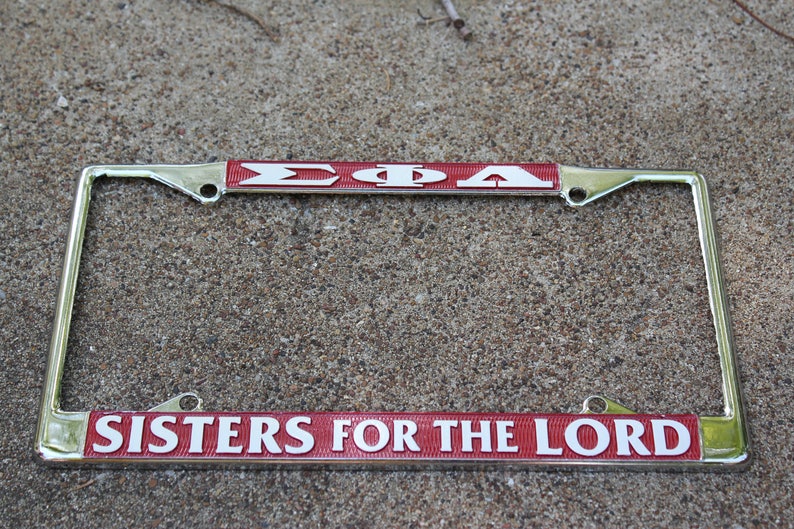 May include: A silver licence plate frame with a red and white band that says "&Sigma;&Phi;&Alpha; SISTERS FOR THE LORD".