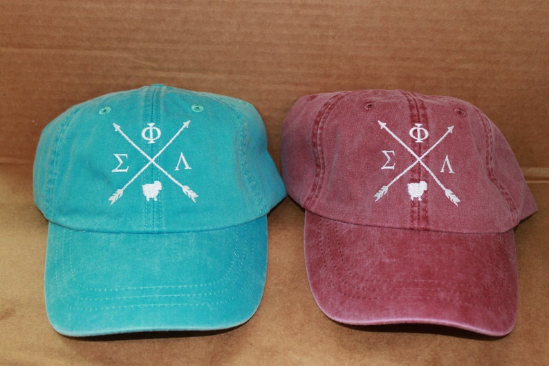 May include: Two adjustable baseball caps with a white embroidered design. The design features a crossed arrow with the letters "&Sigma;&Phi;&Lambda;" and a small sheep. One cap is teal blue and the other is burgundy.