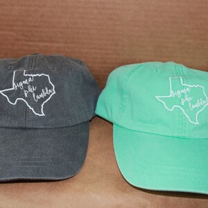 May include: Two baseball caps with an embroidered outline of the state of Texas and the text "Sigma Phi Lambda" inside. One cap is dark gray and the other is light green.