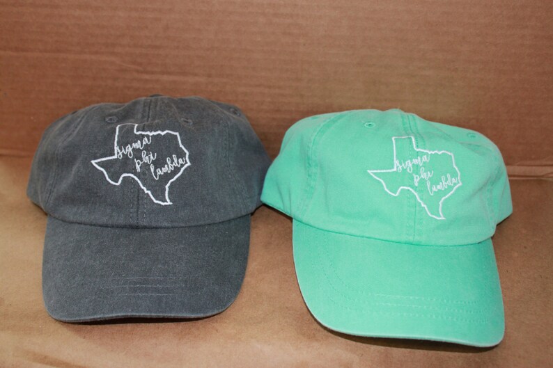 May include: Two baseball caps with an embroidered outline of the state of Texas and the text "Sigma Phi Lambda" inside. One cap is dark gray and the other is light green.