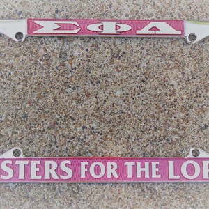 May include: A silver licence plate frame with a pink stripe across the top and bottom. The top stripe says "&Sigma;&Phi;&Alpha;" in white letters. The bottom stripe says "SISTERS FOR THE LORD" in white letters.