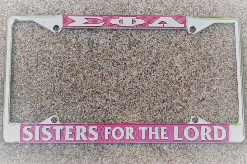 May include: A silver licence plate frame with a pink stripe across the top and bottom. The top stripe says "&Sigma;&Phi;&Alpha;" in white letters. The bottom stripe says "SISTERS FOR THE LORD" in white letters.