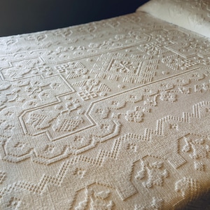 May include: Cream-colored, textured bedspread with an intricate, raised geometric pattern. The design features repeating shapes, including squares, rectangles, and floral motifs. The bedspread is neatly arranged on a bed, with the pillow visible at the top.