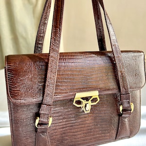 May include: A brown leather handbag with a gold clasp and buckle closures. The bag has a textured, embossed pattern that resembles snakeskin.