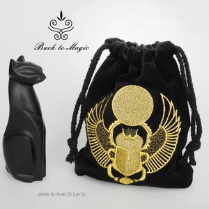 May include: A black cat figurine and a black velvet drawstring bag. The bag features a gold embroidered design of a sun disc with wings and a scarab beetle. The text "Back to Magic" is above the cat.