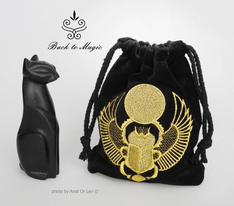 May include: A black cat figurine and a black velvet drawstring bag. The bag features a gold embroidered design of a sun disc with wings and a scarab beetle. The text "Back to Magic" is above the cat.