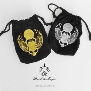 May include: Two black velvet drawstring bags with embroidered scarab beetle designs. One bag features a gold beetle, the other a silver beetle. The bags have black drawstrings. The text "Back to Magic" is below the bags.