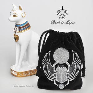 May include: A white cat statue with gold accents and an ornate collar stands next to a black drawstring bag. The bag features a silver embroidered scarab beetle design. The text "Back to Magic" is visible.