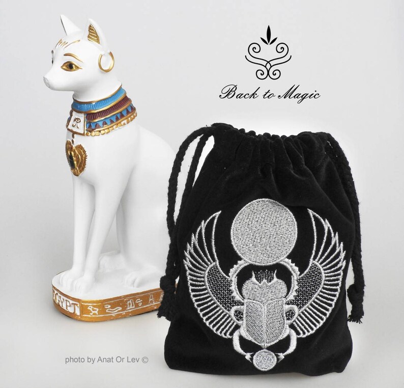 May include: A white cat statue with gold accents and an ornate collar stands next to a black drawstring bag. The bag features a silver embroidered scarab beetle design. The text "Back to Magic" is visible.
