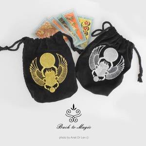 May include: Two black velvet drawstring bags with embroidered scarab beetle designs, one gold and one silver. Tarot cards are visible behind the bags. The bags have a decorative design and the text "Back to Magic".