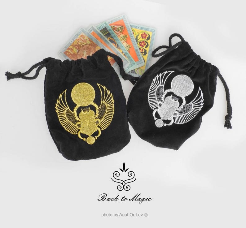 May include: Two black velvet drawstring bags with embroidered scarab beetle designs, one gold and one silver. Tarot cards are visible behind the bags. The bags have a decorative design and the text "Back to Magic".
