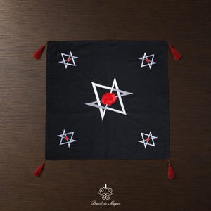 Pode incluir: Um pano de tecido preto quadrado com um grande contorno de estrela prateada no centro e uma rosa vermelha. Quatro contornos de estrelas prateadas menores estão posicionados nos cantos, cada um com um ponto vermelho. Borlas vermelhas nos cantos.