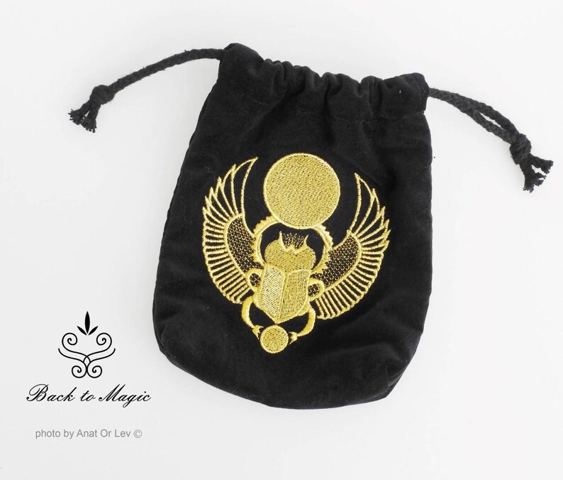 May include: Black velvet drawstring bag with a gold embroidered scarab beetle design. The bag features a gold sun disc above the beetle and wings. The bag is labeled "Back to Magic".