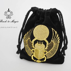 May include: Black velvet drawstring bag with gold embroidered scarab beetle design. The bag features a gold sun and wings, and the text "Back to Magic". The bag is closed with a black rope.