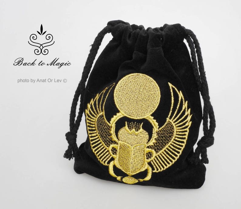 May include: Black velvet drawstring bag with gold embroidered scarab beetle design. The bag features a gold sun and wings, and the text "Back to Magic". The bag is closed with a black rope.