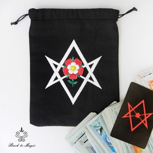 Op de afbeelding: Een zwarte trekkoordtas met een wit geborduurd pentagram en een rode roos in het midden. De tas ligt op een wit oppervlak met een tarotkaartendek op de achtergrond.