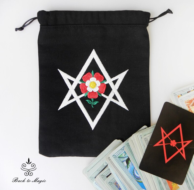 Op de afbeelding: Een zwarte trekkoordtas met een wit geborduurd pentagram en een rode roos in het midden. De tas ligt op een wit oppervlak met een tarotkaartendek op de achtergrond.