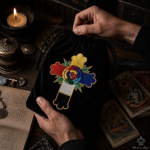 May include: A black velvet drawstring pouch featuring an embroidered cross design. The cross is composed of red, yellow, blue, and white segments with a central circular rainbow-colored floral motif and green starburst accents at the center.