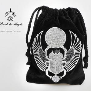 May include: Black velvet drawstring bag with a white embroidered scarab beetle design. The bag features a detailed depiction of the beetle with wings, a sun disc, and other Egyptian symbols. The text "Back to Magic" is also visible.