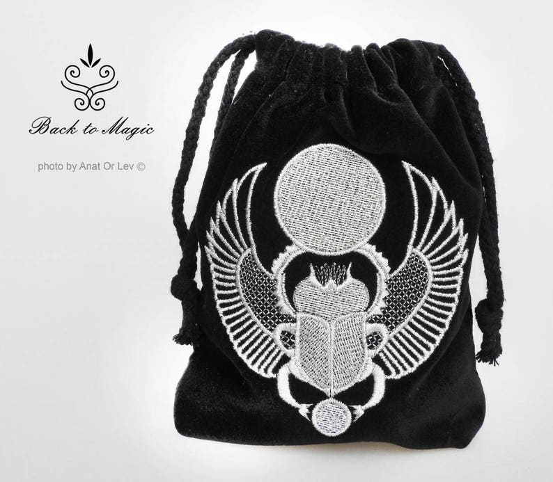 May include: Black velvet drawstring bag with a white embroidered scarab beetle design. The bag features a detailed depiction of the beetle with wings, a sun disc, and other Egyptian symbols. The text "Back to Magic" is also visible.