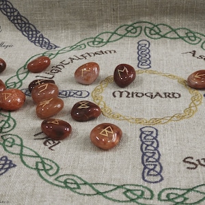 Könnte beinhalten: Ein Set aus 15 Karneol-Runensteinen mit goldenen Lettern auf einem Leinenstoff mit einem gestickten Symbol der neun nordischen Reiche. Die Runen sind in einem Kreis um das Symbol angeordnet. Der Stoff trägt die gestickten Worte "Back to Magic" und "Midgard".