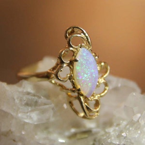 May include: A gold ring with a white opal gemstone set in a delicate, ornate design. The ring is set on a bed of white crystals.