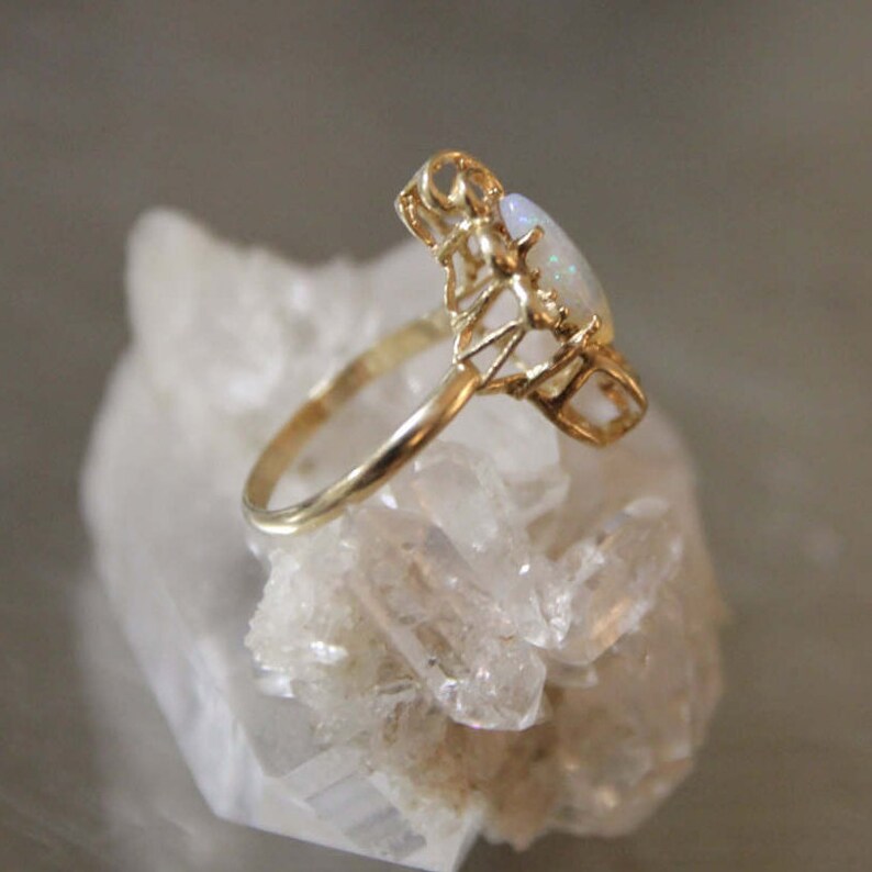 May include: A gold ring with a white opal gemstone set in a delicate, intricate design. The ring is on a bed of white crystals.
